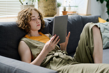 Young relaxed Caucasian woman in casualwear looking through online data on screen of digital tablet...