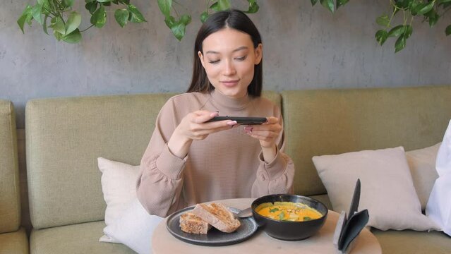 Young asian woman takes picture of lunch on phone in cafe. Lady has vegetable pumpkin soup with pieces of grain bread. Blogger sits on comfortable couch