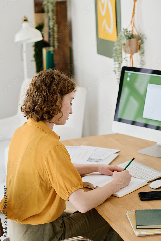Aufkleber Young businesswoman in casualwear making notes in notebook while sitting in front of computer monitor and planning work
