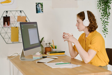 Young serious female freelancer with cup of tea or coffee looking at computer screen with small...