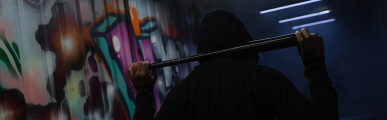 Back view of african american bandit in hoodie holding baseball bat in garage with graffiti, banner
