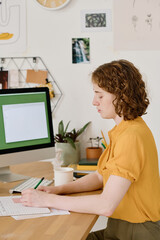 Fototapeta premium Serious girl in casualwear drawing sketch or scheme of new project in copybook while sitting in front of computer monitor by workplace