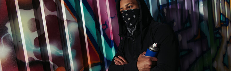 African american vandal with covered face holding spray paint near graffiti on urban street, banner