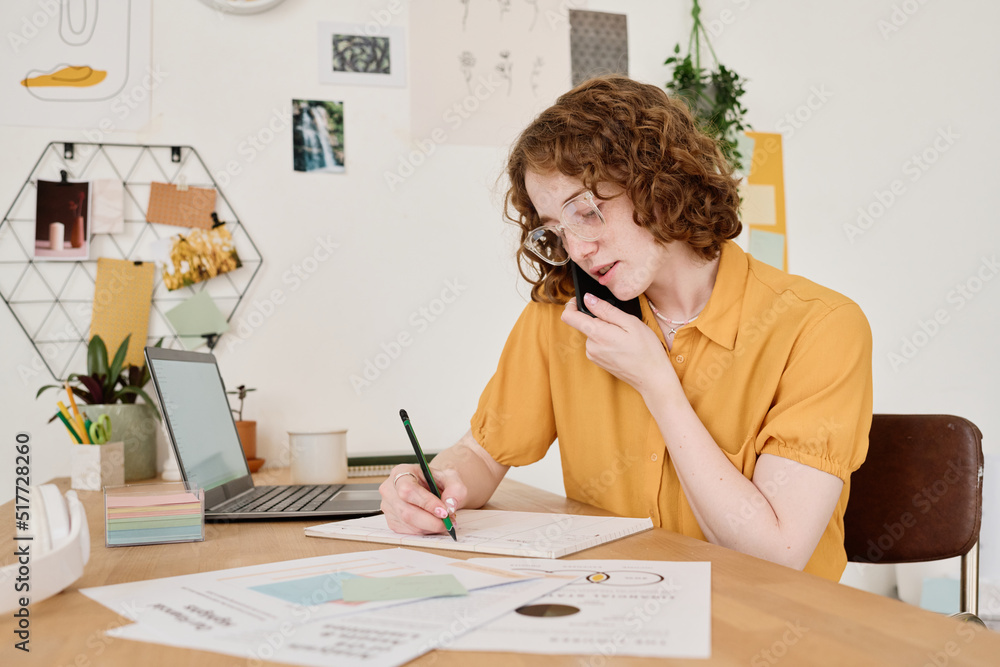 Poster young female freelancer consulting clients on mobile phone and making notes on paper while analyzing