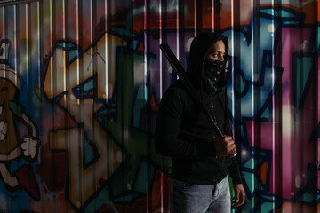 African american vandal with mask on face holding baseball bat near graffiti on urban street