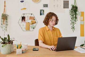 Young student or freelancer in casualwear sitting in front of laptop by workplace at home and looking through online data on screen