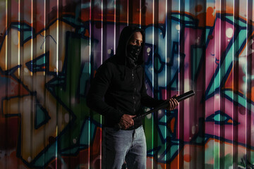 African american vandal in hoodie holding baseball bat near graffiti outdoors at night