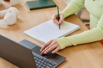 Young female student making notes in copybook while looking through online data during work over new school project in front of laptop