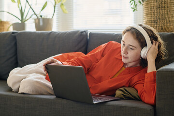 Youthful girl in headphones lying on soft comfortable couch in living room and looking at laptop screen while watching online training