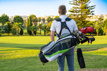 Back view of young handsome man at the course carrying a golf bag.