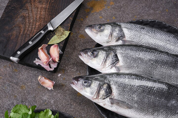 Fresh raw fish Seabass on a black plate with spices. Three fresh fishes with garlic, bay leaf and...