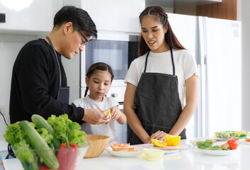 Happy family cooking together in the kitchen. Father, mother, and cute little daughter turn vegetables to make salads