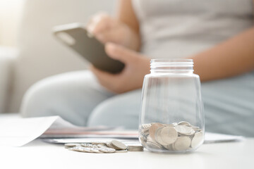 Savings and calculating budget concept with woman, mobile phone, and coin jar in a living room.