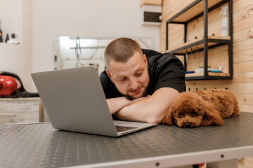 Professional male groomer working on laptop in her workplace in grooming salon near with poodle dog. Animals Grooming concept