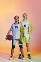 Fototapeta premium Two sportive girls, teens, basketball players standing together with basketball ball isolated on pink background in neon light. Concept of sport, team, enegry, competition, skills
