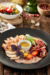 Meat platter on black plate on wooden background. Meat assorted with sauce and basil. Composition with Italian antipasti in rustic style. Meat delicatessen on dark wooden table with ingredients.