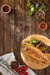 Grilled rainbow trout with herbs on wooden background. Composition with roasted rainbow trout and ingredients. Grilled whole fish on wooden table in rustic style.