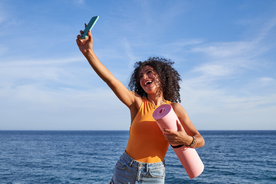 Cheerful Hispanic woman with mat taking selfie on seashore