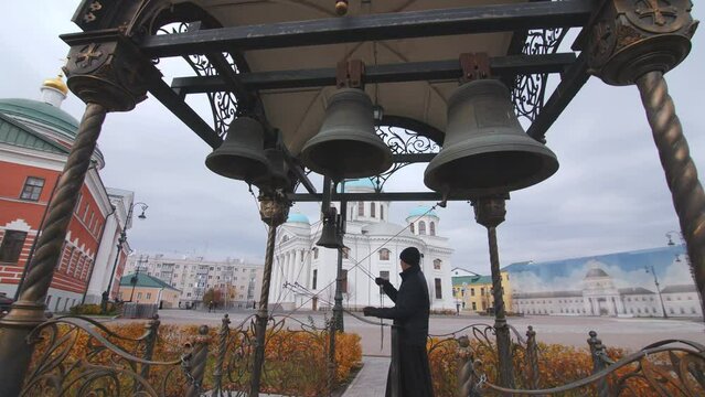Orthodox monk rings large church bells in belfry at ancient christian monastery on gloomy day. Religion service