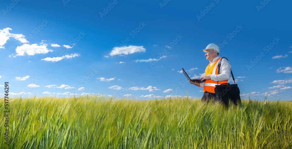 Poster a man in a helmet and an orange vest walking along the field with grass. engineer builder examines t