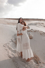 Fototapeta na wymiar female energy rustic party. two happy brunette girls in white boho style dresses are standing close in hugs and laughing on the sand career background in desert. esoteric lifestyle concept, free space