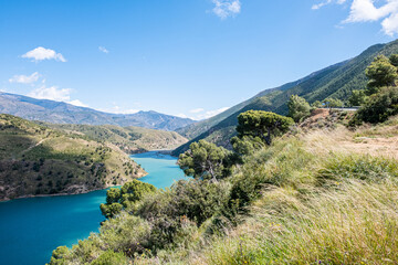 Fototapeta na wymiar Breath-taking view from the top on green lake surrounded by picturesque mountain alley with trees and bushes in sunny summer day with blue sky and white clouds in background.