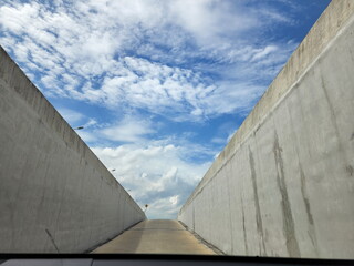 A view from inside the vehicle as it moves out of the concrete tunnel into the road. You can see the bright blue sky and white clouds scattered across the sky.
