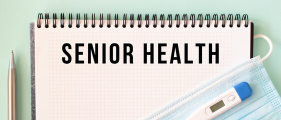 A medical mask and a thermometer are on the notepad. SENIOR HEALTH text in a notebook.