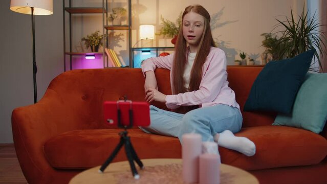 Young redhead child girl blogger taking selfie on smartphone tripod, communicating video call online with subscribers. Teen freckles kid children at modern home apartment living room sitting on couch