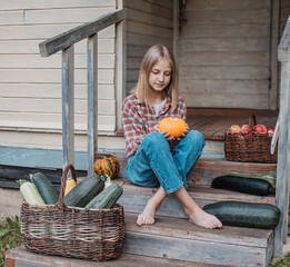 Girl is sitting on steps of an old village house with pumpkin in hands near baskets with zucchini and apples. Harvest. Autumn. Halloween