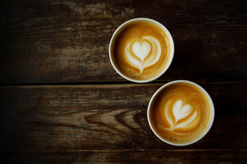 Two disposable cups of coffee with the heart on foam on a wooden table.