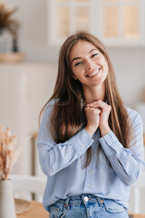 Vertical shoot of grateful European girl with long loose hair is standing in the kitchen in a blue shirt and jeans, looking at the camera, satisfied woman toothy smiling folded her arms on her chest.