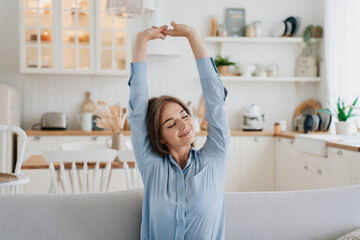 Gorgeous blonde caucasian young woman in blue shirt sitting eyes closed on couch stretching  rising up hands smiling. Pretty swedish girl  relaxing at home against kitchen. Comfort and cozy home.