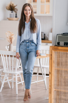 Vertical shot of beautiful girls in jeans and a blue shirt standing in the kitchen, looking at the camera, smiling contentedly. Beautiful satisfied european blonde housewife home relaxing on weekend.