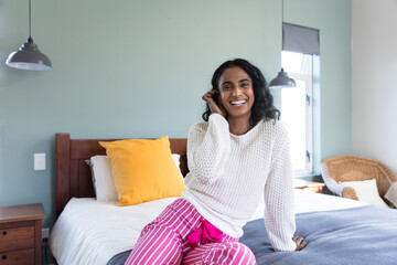 Portrait of cheerful biracial young woman smiling while relaxing on bed at home, copy space