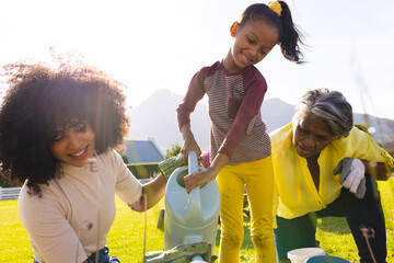 Multiracial happy girl with mother and grandmother watering plants against clear sky in yard