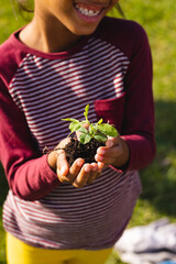 Midsection of biracial smiling girl holding compost and sapling while standing in yard