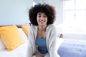 Fototapeta na wymiar Portrait of smiling biracial young woman with afro hair wearing jacket relaxing on bed at home