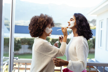 Side view of biracial female friends applying beauty cream on each others faces in balcony