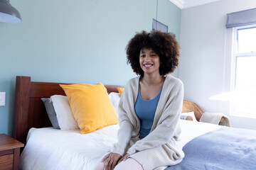 Portrait of smiling biracial young woman with afro hair wearing jacket sitting on bed at home