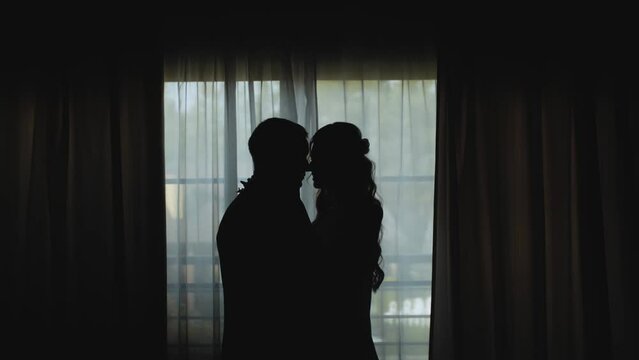 Silhouette of newlywed, loving couple standing near the window. Bride kiss the groom slow motion.