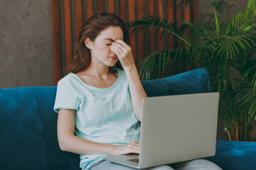 Young woman she 20s in casual clothes mint t-shirt keep eyes closed rub put hand on nose hold use work on laptop pc computer study online sit on blue sofa indoor rest at home in own room apartment