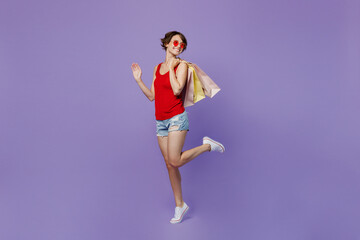 Full body side view young smiling happy woman 20s she wears red tank shirt eyeglasses holding package bags with purchases after shopping look aside on wotkspace area isolated on plain purple backround