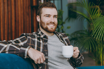 Young dreamful man he wears brown shirt hold cup of coffee look aside in reverie good mood sitting on blue sofa in own living room apartment stay home indoor flat on weekends People lifestyle concept