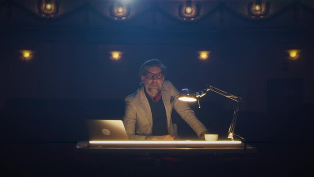 The director in glasses and in a suit sitting at a table in a dark theater and thoughtfully watching the rehearsal of the play
