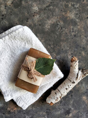 Handmade tar soap in craft paper tied with twine on a white terry towel with birch bark, leaf on a black concrete background with copy space. Bath concept.