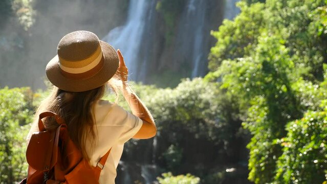 Back view of woman with photo camera shooting tropical waterfall. Inspiring millennial travel blogger in journey. Happy young traveler girl on summer vacation. Concept of wanderlust and adventure