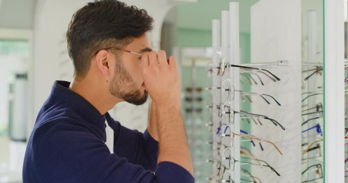 Young man choosing glasses in optometry clinic. Patient trying and testing eyewear frames for optometrist prescription lenses. Man using mirror to admire reflection with new vision wear from optician