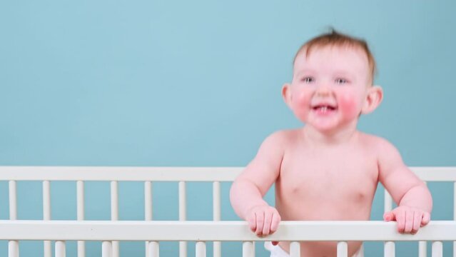 Happy infant baby boy stands in the crib, studio blue background. Smiling child in white diaper
