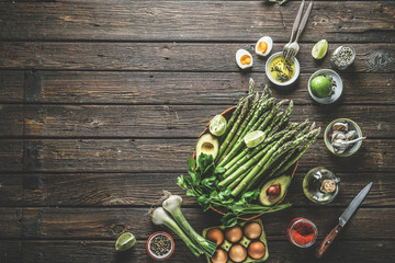Food background with green asparagus and various healthy ingredients: avocado, egg, spring onion, lime, spices and herbs at dark rustic wooden kitchen table. Tasty cooking at home.Top view.